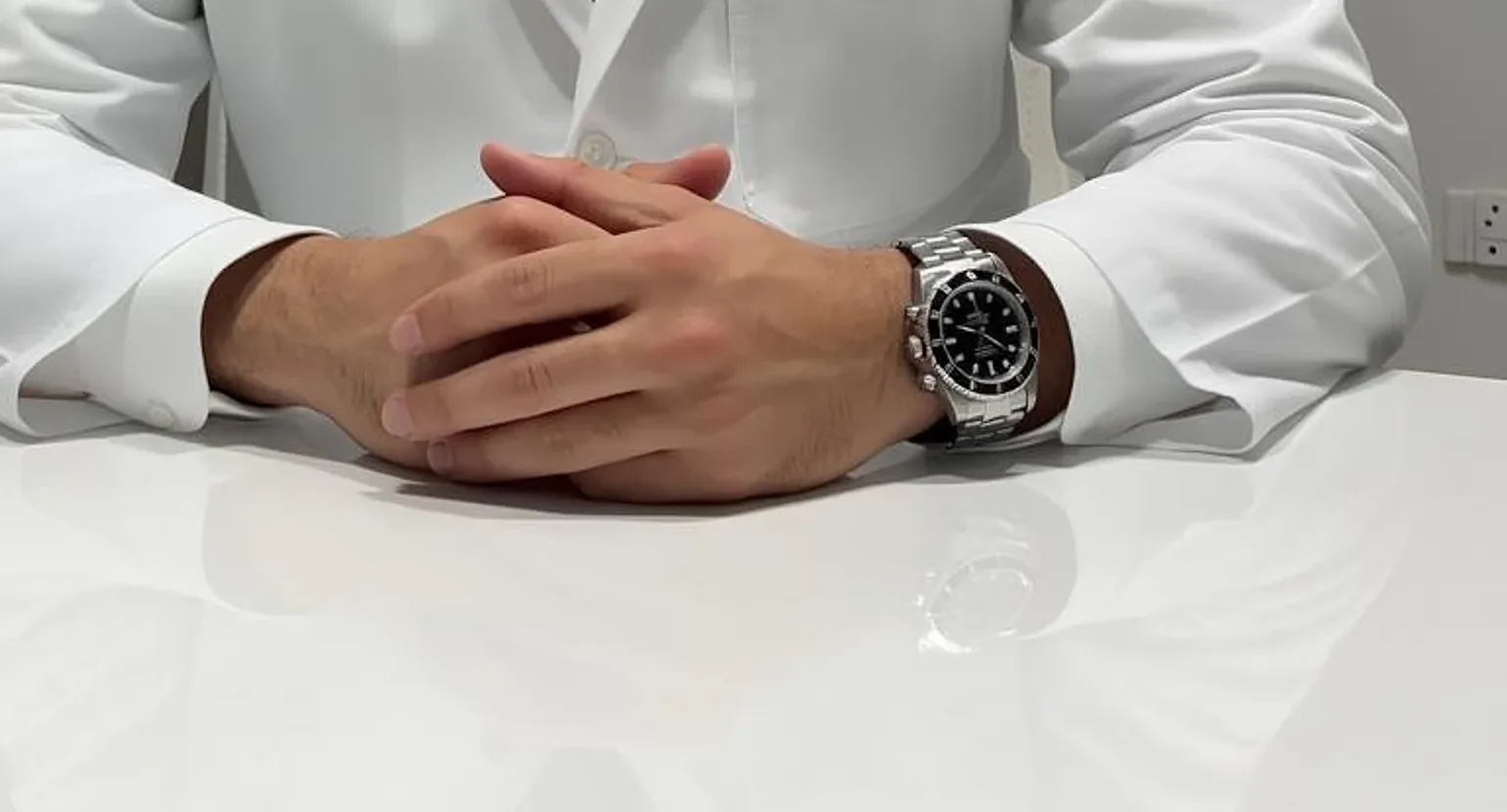 As mãos do Dr. Thiago cruzadas sobre a mesa branca do consultório, de jaleco